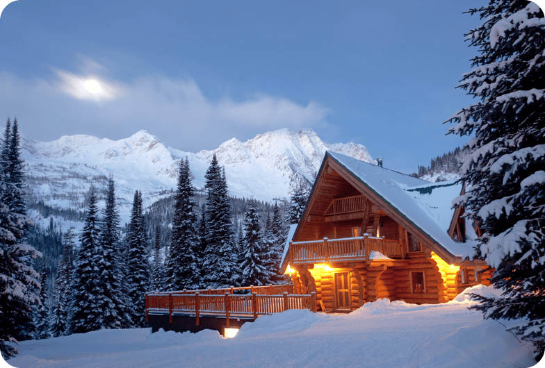 Outside view of a log cabin with the roof covered in snow, trees, and a snow covered mountain in the background.
